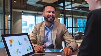 Homem de terno conversando com outra pessoa e um notebook aberto com gráficos