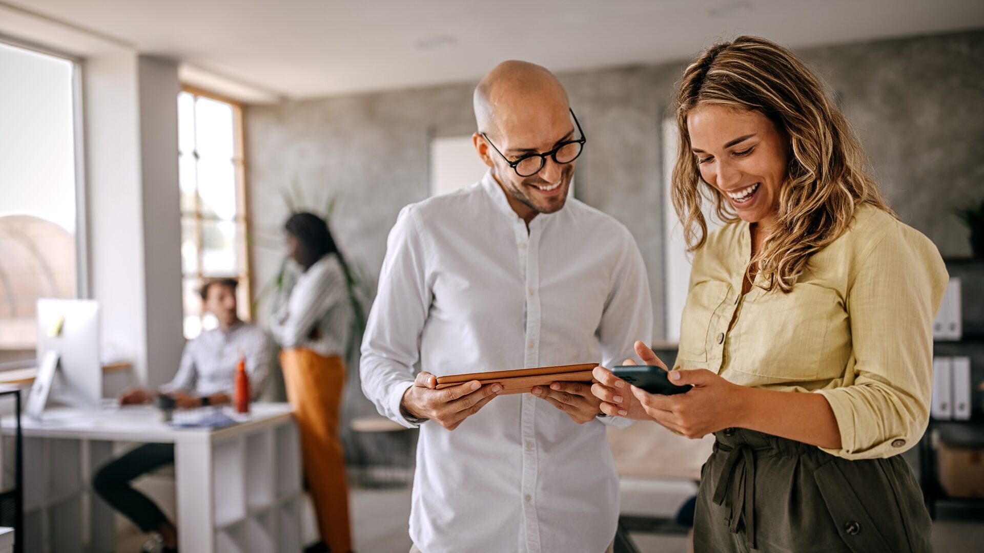 Duas pessoas sorrindo em um escritório com tablet e celular nas mãos